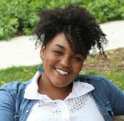 Outdoor photo of a smiling Black Woman early middle age in a button down white shirt and a Blue cardigan
