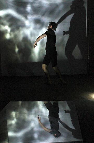 Actor, Will Swyers, in twisting pose in front of tall projection of water ripples reflecting down into a pool of water on the stage floor