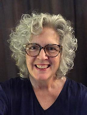 Juanita Rockwell photo showing a woman in her 60s with curly white hair, fair skin and dark rimmed glasses. She is smiling broadly and wearing a black V-neck shirt.