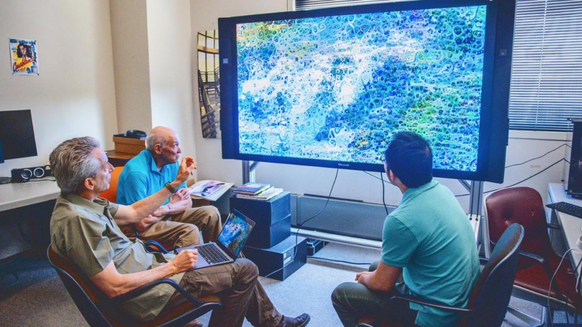 A color photo showing 3 men looking at artwork on a large digital display