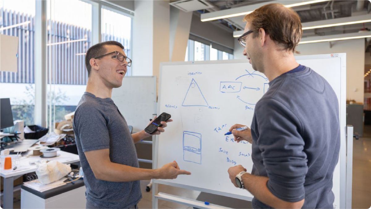 A color photo of 2 men discussing a project in front of a white board.