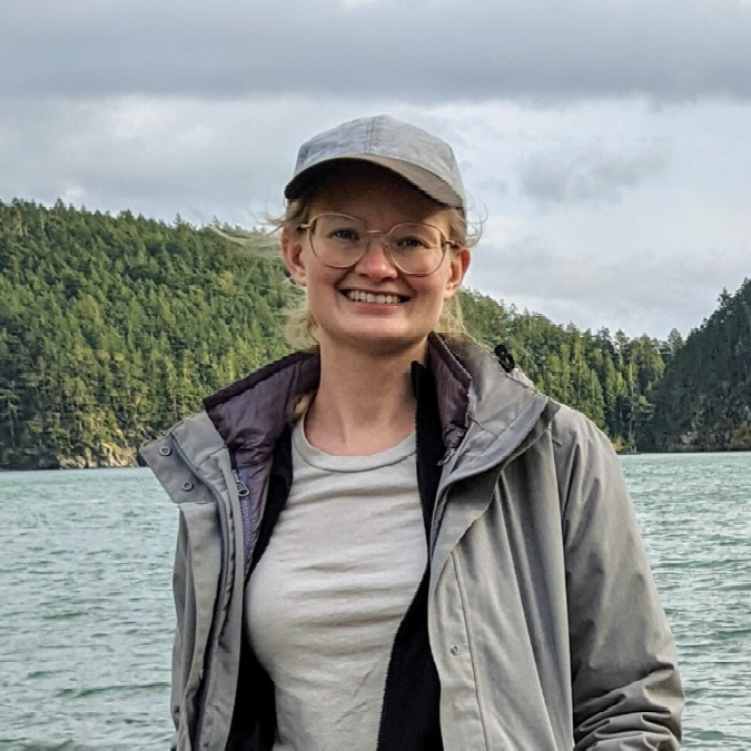 A color photo of a woman smiling for a photo in front of a lake.
