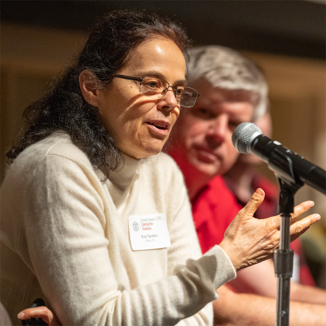 A color photo of a woman speaking at a microphone with a man in the background listening