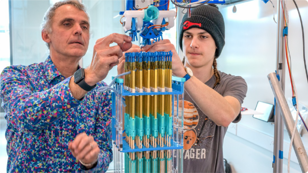 A color photo showing 2 men working on a knitting device.