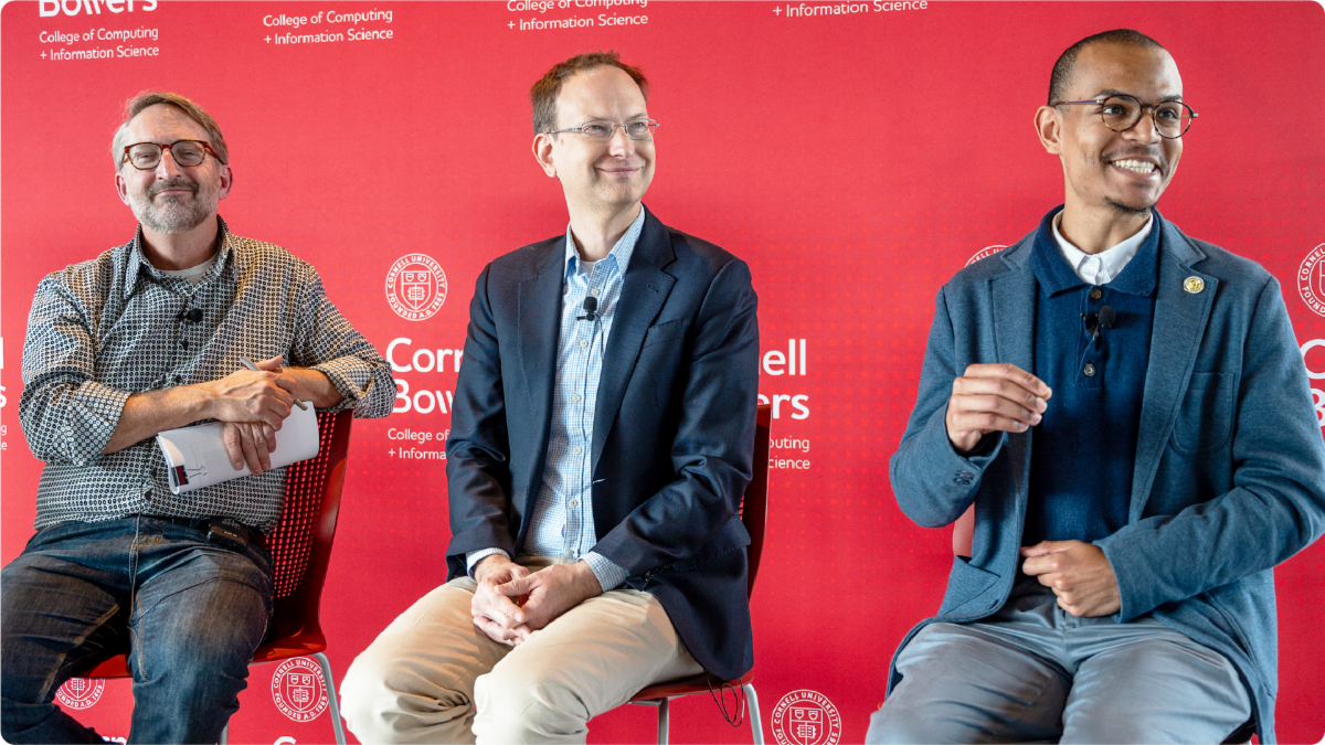A color photo showing 3 men sitting on a stools on a stage.