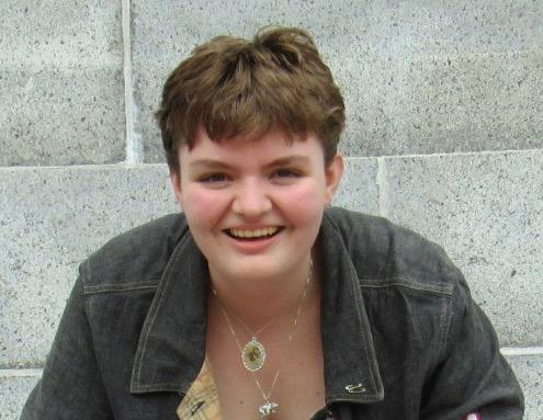 A light skinned nonbinary individual in their 20s with short brown hair smiles wearing a dark jacket and layered necklaces in front of a light gray stone wall.
