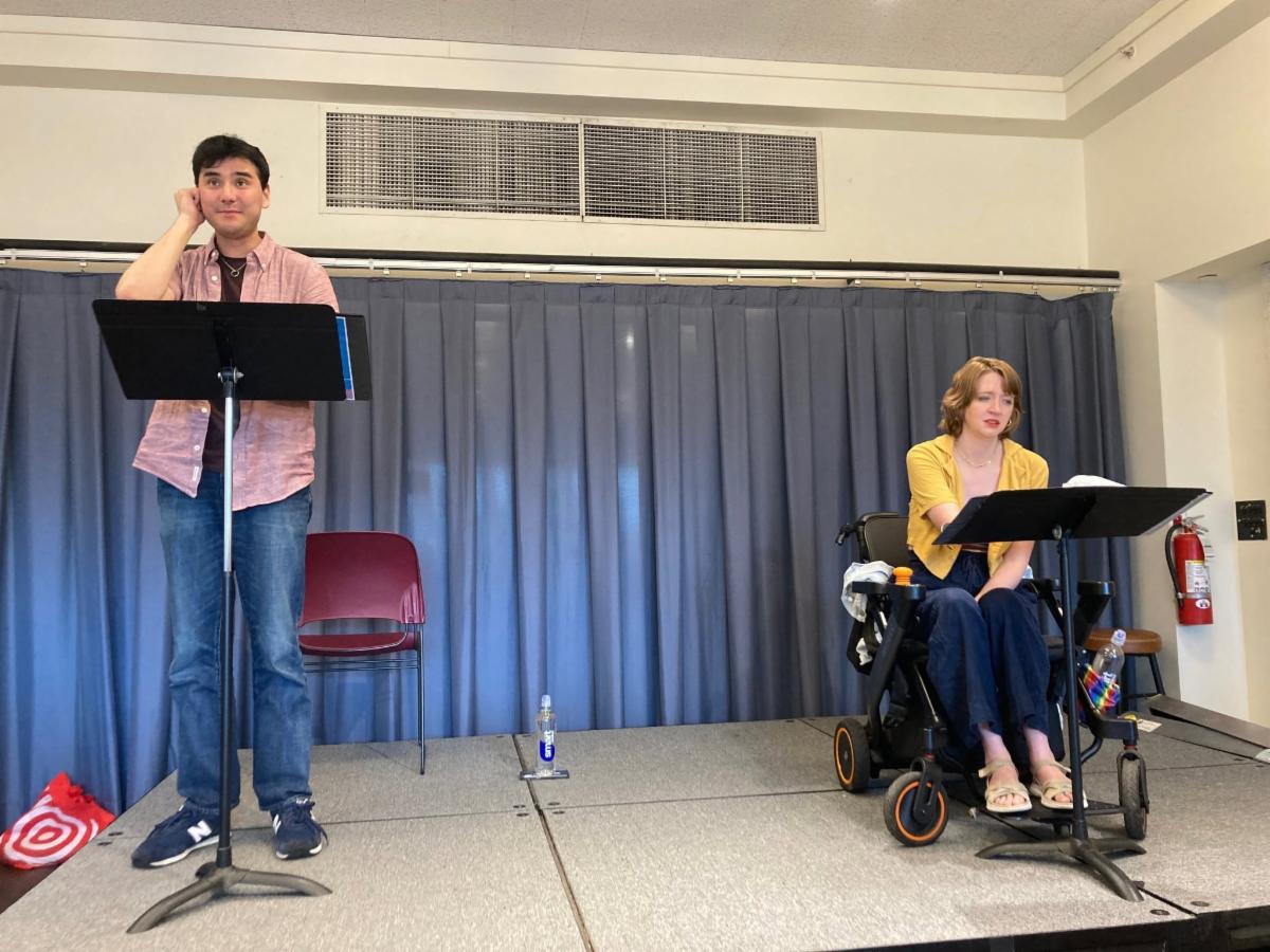 2 light skinned actors a man with black hair and a woman with strawberry blonde hair. The man stands and acts like he is a phone on a stage behind a music stand. The woman is in a wheelchair behind a music stand. They are in casual clothes and engaged in their scripts.