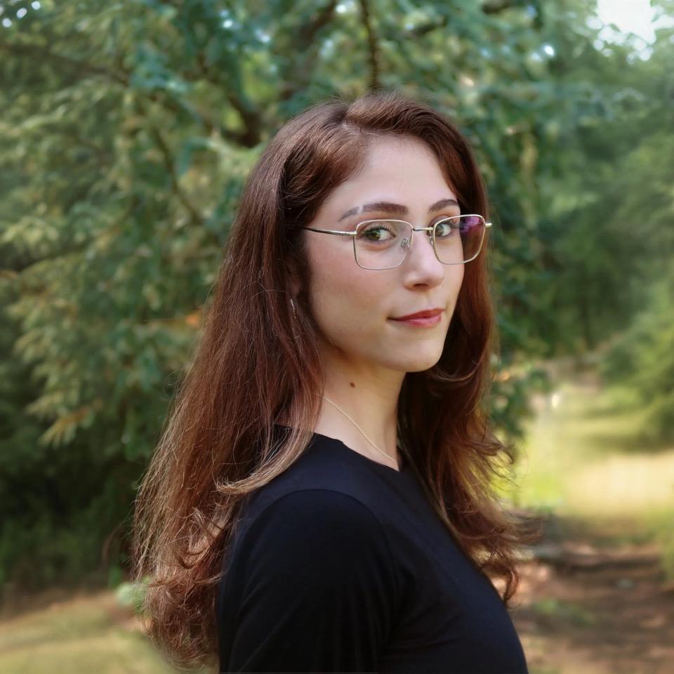  Abrianna Sigel a light-skinned woman 20s long brown hair glasses wearing a black shirt standing outdoors with a large tree behind her.