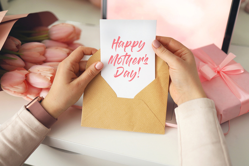 Woman s hands holding a letter in craft envelope. Pink background_ Mother s day concept. Tulips flower and pink gift box in background. Womens home desk.