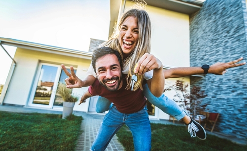 Happy young couple holding home keys after buying real estate - Husband and wife standing outside in front of their new house