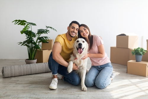 Cheery young international couple with cute golden retriever dog sitting on floor of new home on relocation day. Happy millennial husband and wife with cute pet moving to new house