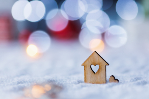 Wooden icon of house with hole in form of heart with red home Christmas decor and blurred bokeh background in daylight. New Year card with copy space. Concept of sweet home.
