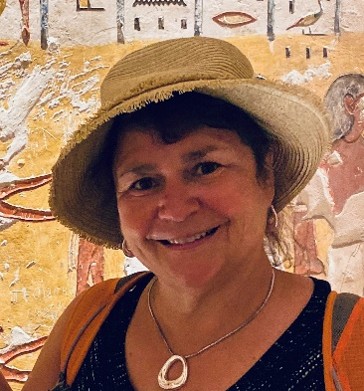 Photo of OLLI instructor Janinne Johnson smiling and wearing a hat while standing in front of hieroglyphs