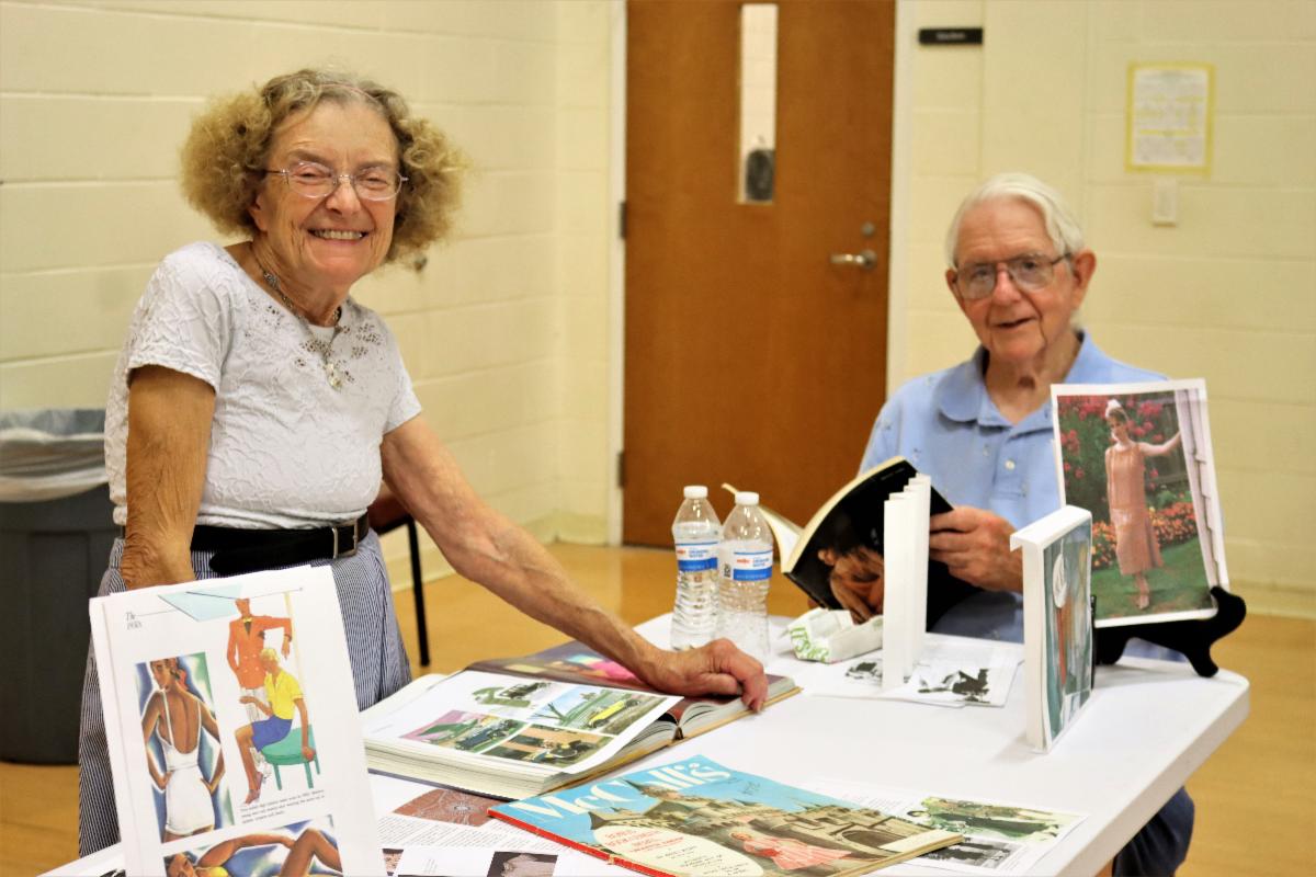 2 senior aged adults, a smiling woman in a dress on the left, and a man sitting down on the right with a book open. Both have a table setup advertising their course on Fashions from the Past, during an OLLI at UK Open House at a church gymnasium.
