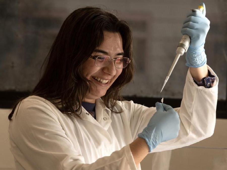 Researcher in a lab coat and gloves uses a pipette to transfer liquid into a small tube, smiling while working at a lab bench.