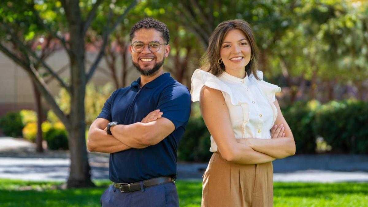 A man and a woman stand with their arms crossed smiling outside