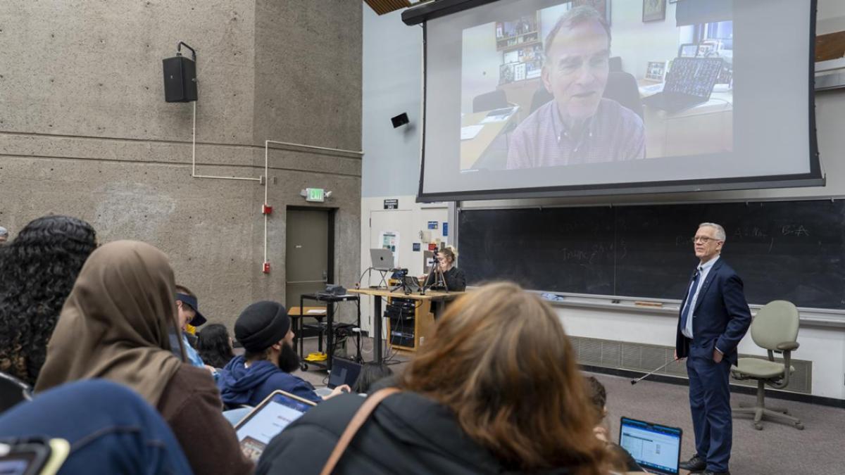 Lecturer stands at the front of a classroom as students watch a large screen showing a guest speaker on a video call.