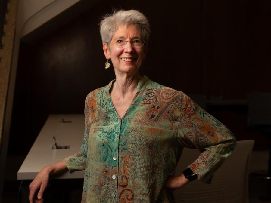 Smiling person with short gray hair glasses and patterned shirt standing indoors with hand on hip