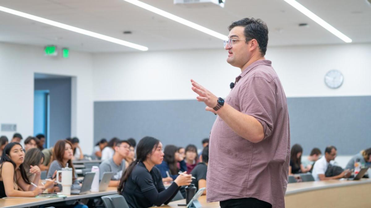 Man stands speaking at the front of a classroom while students sit at desks listening and taking notes