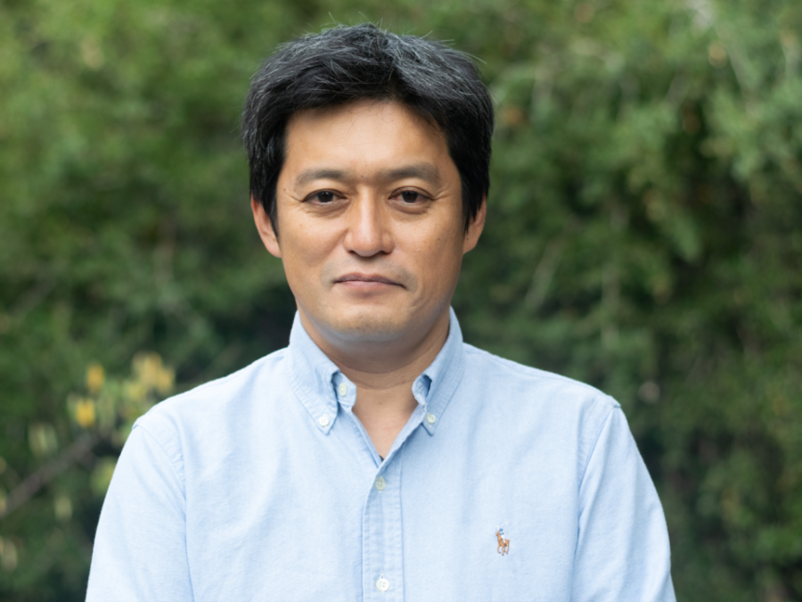 A man in a light blue shirt standing outside in front of foliage