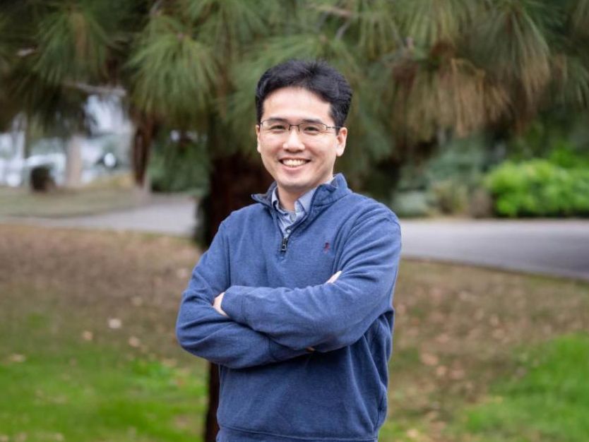 A man wearing glasses and a blue pullover stands outdoors with his arms crossed smiling at the camera