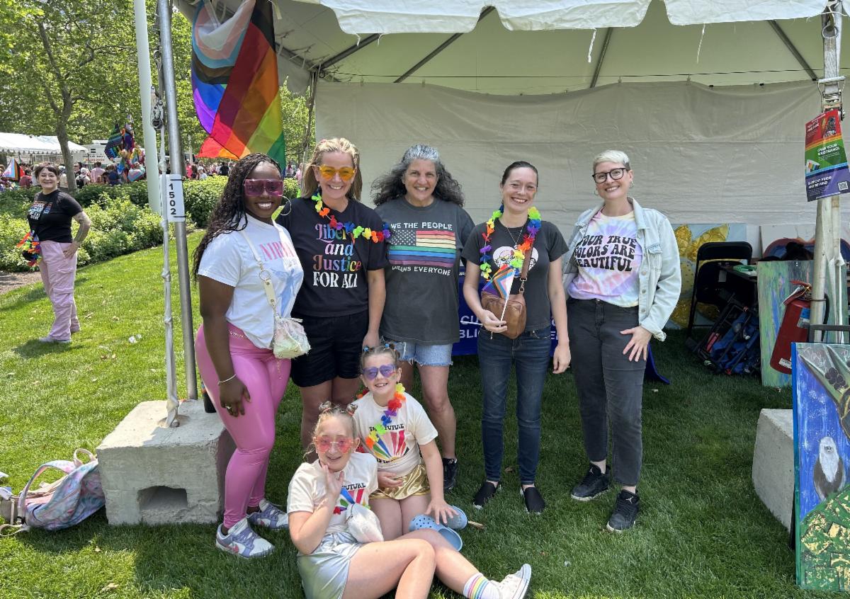 five people stand near a pride tent with two youth sitting on the ground in front