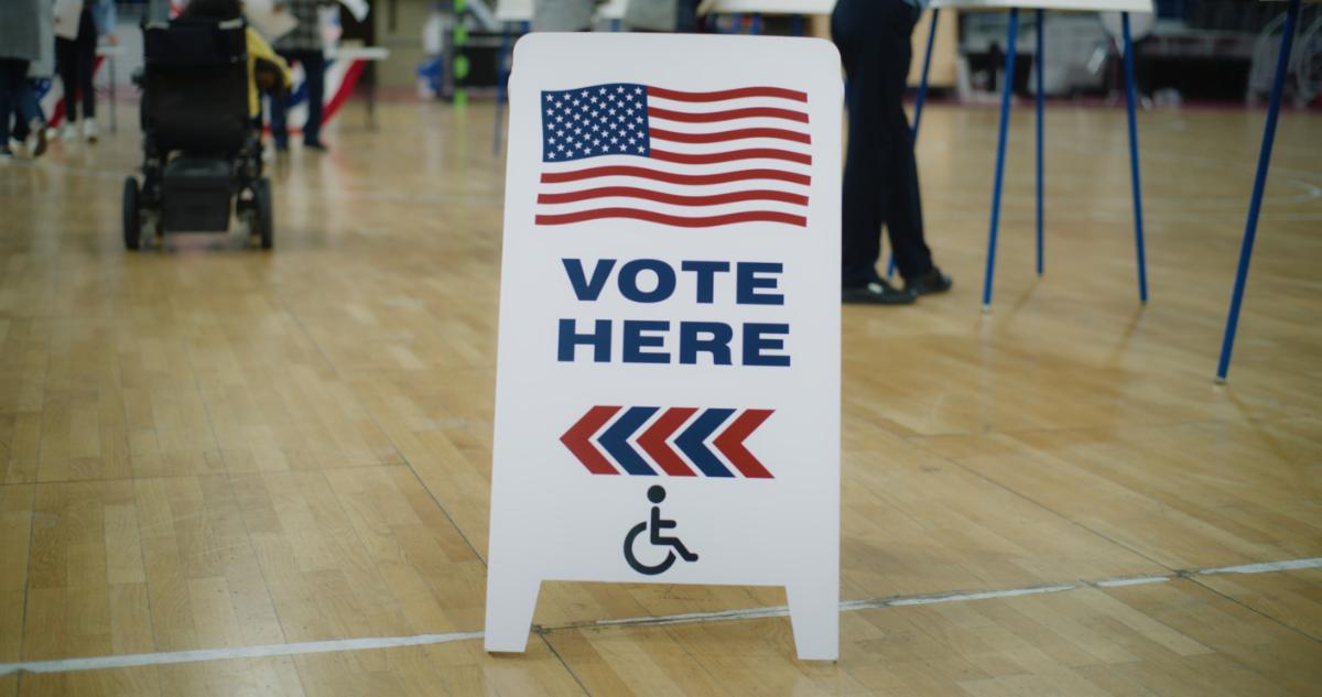 Inside a school gym that serves as a polling place, a sign with an American flag and a wheelchair icon says vote here.