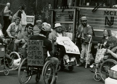 Black and white photo of Greyhound protest in 1997. Man in wheelchair with sign on back that says I can't even get to the back of the bus