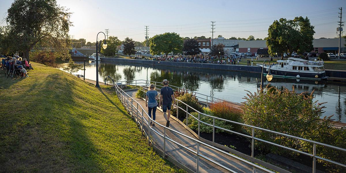 Two people walk down to the Erie Canal at sunset in Newark as a concert is about to begin on the opposite shore