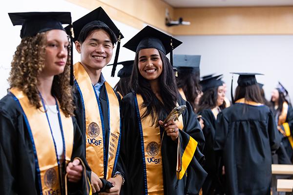 Three students in their caps and gowns and more gathered behind them at graduation