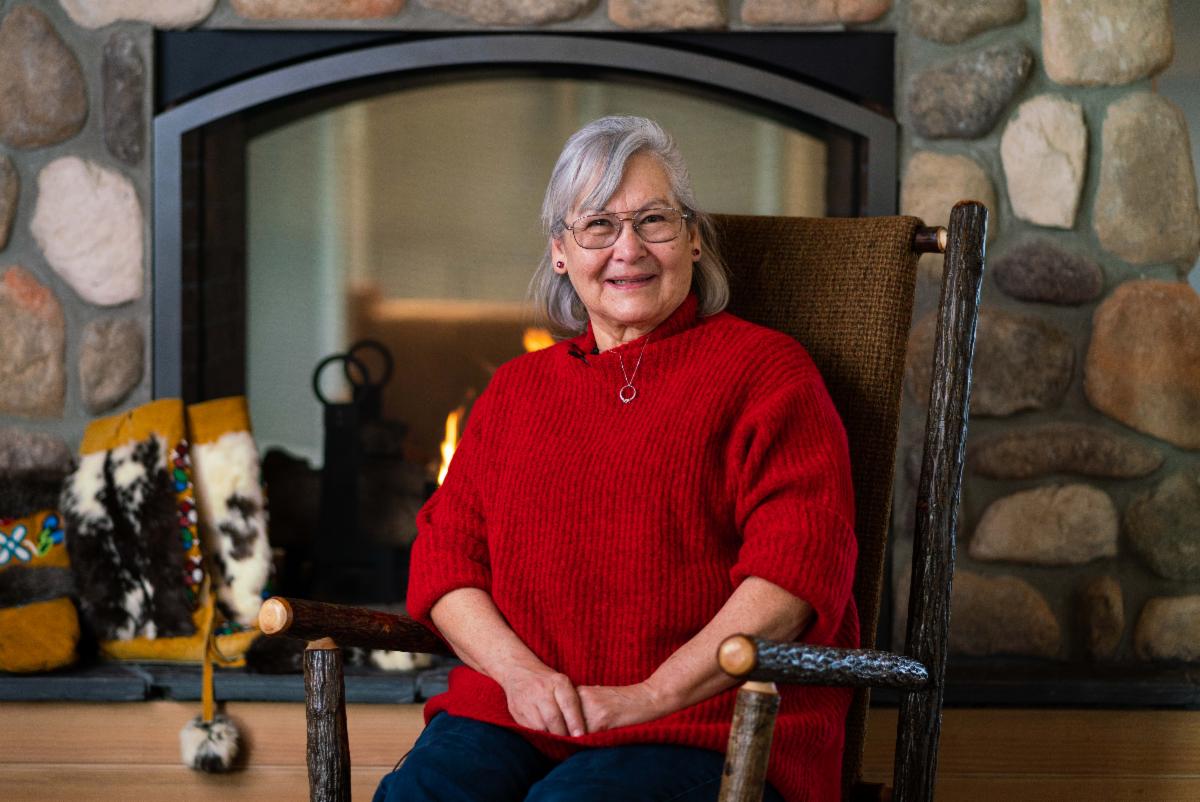 Woman with grey hair, red glasses, red sweater seated by fire place smiling, Lilyrose Meyers