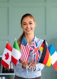 Happy female tutor showing bunch of diverse flags cheerfully smiling at camera_ friendly teacher recommending foreign language studying school_ posing in office