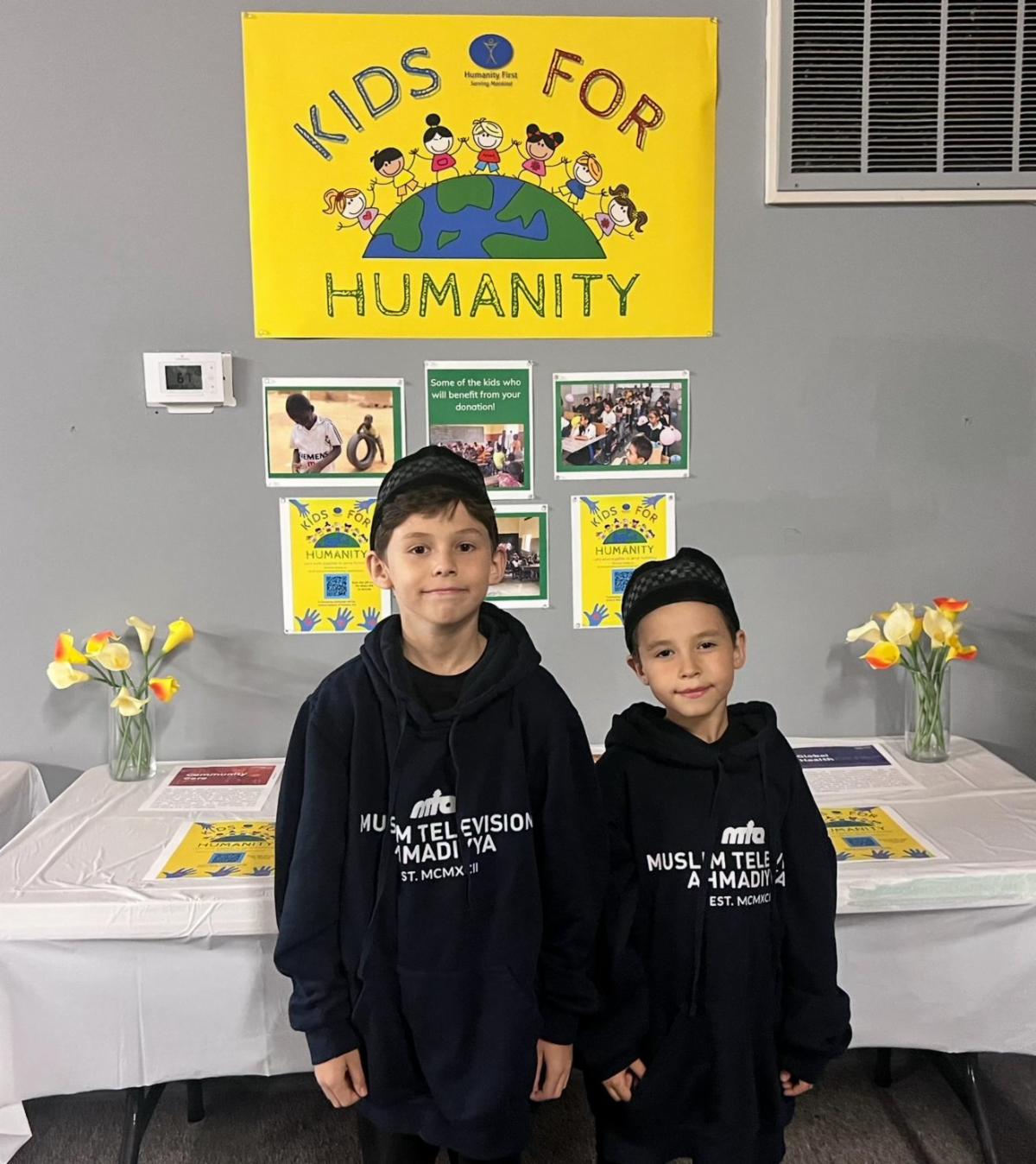 Two young boys in matching black sweatshirts stand in front of a sign kids for humanity and a table with informational brochures