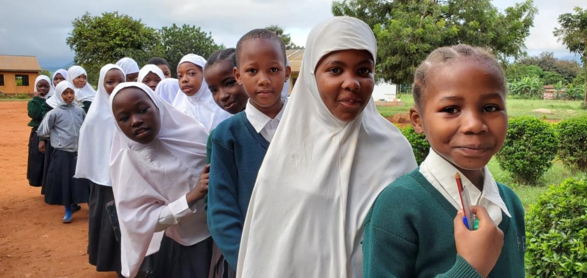 Young girls stand in line wearing school uniforms of white collared shirts and green sweaters. some girls wear a white hijab.