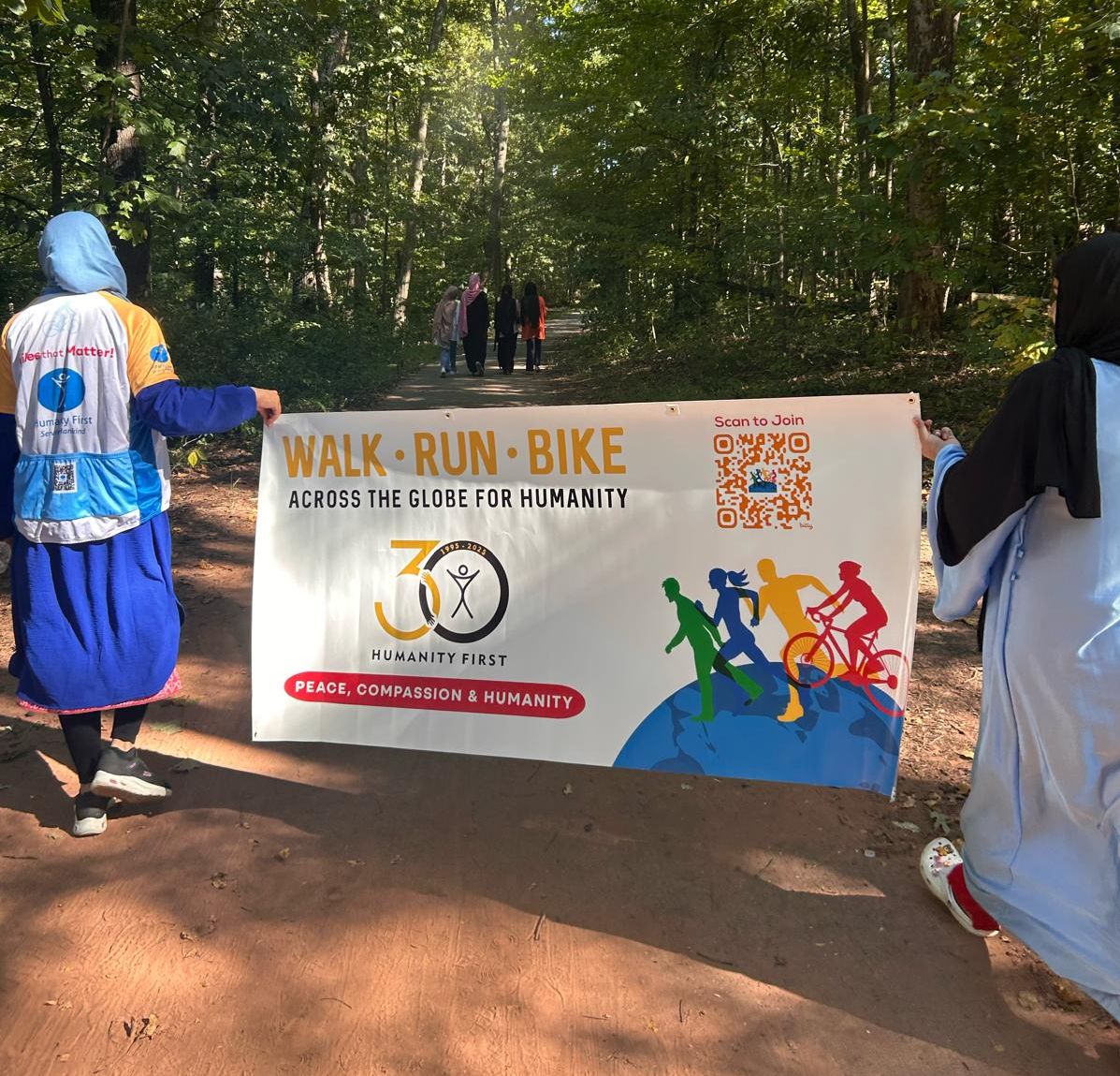 Two ladies in long clothes one of whom wears a humanity first cycling jersey over her clothes carry a large banner for the humanity first walk run bike event on a wide path through the woods