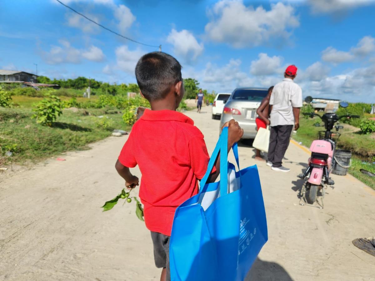 A child in a red shirt walks away from the camera on a dirt road under a serene blue sky. He carries a blue canvas back with the humanity first logo on it.