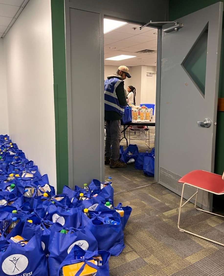Through an open door a person wearing a humanity first blue vest is seen packing bags while dozens of blue bags full of items line the hallway.