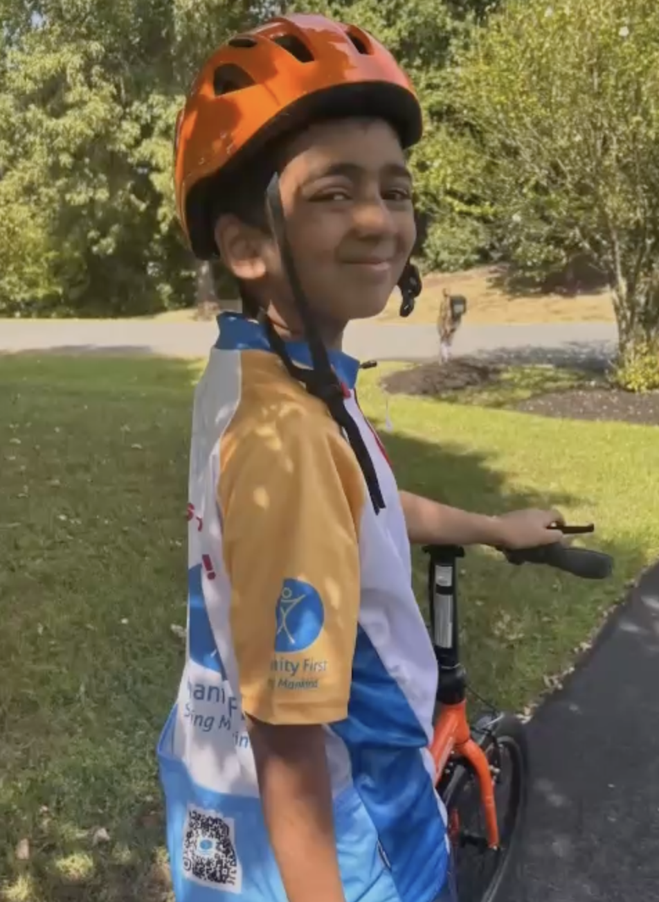 A young kid wearing a humanity first cycling jersey and an orange helmet smiles for the camera