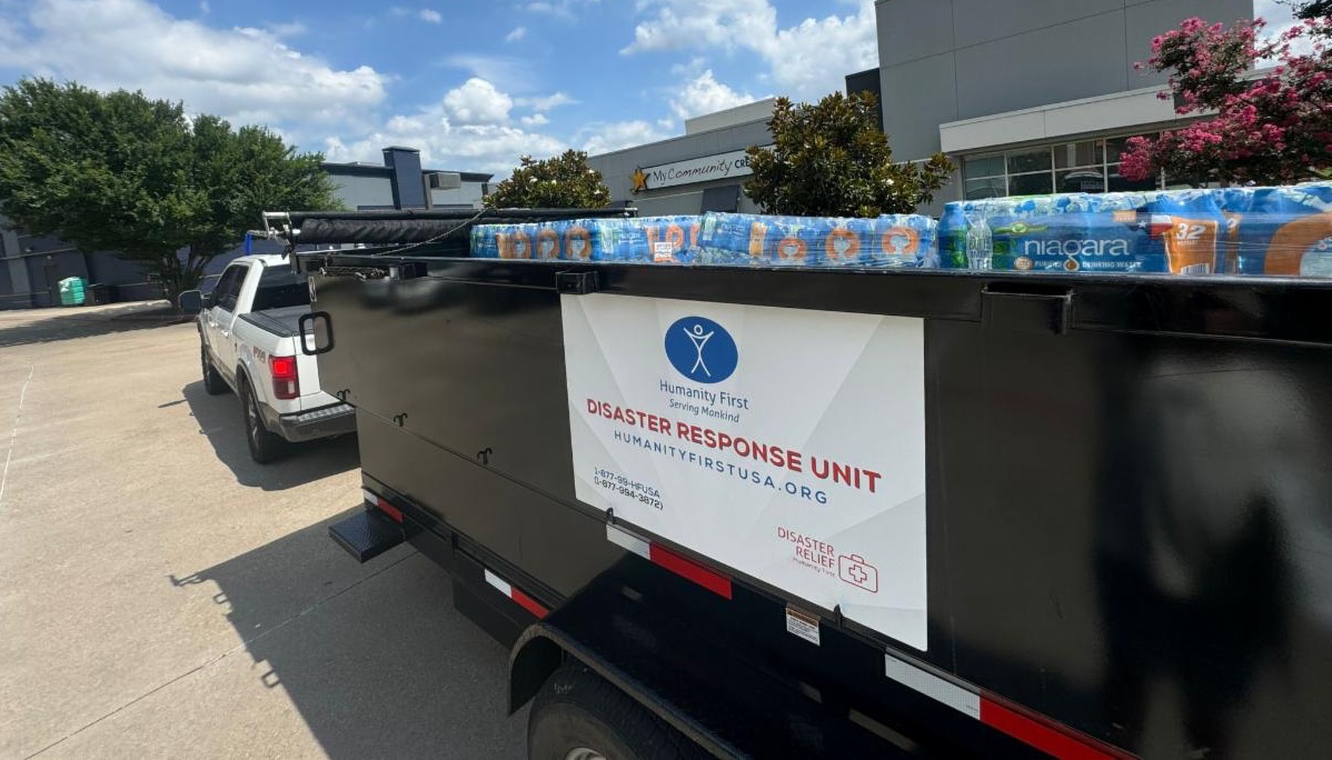 A white pick up pulls a large open bed trailer that is packed with pallets of plastic water bottles and the side bears a decal for the humanity first disaster response unit.
