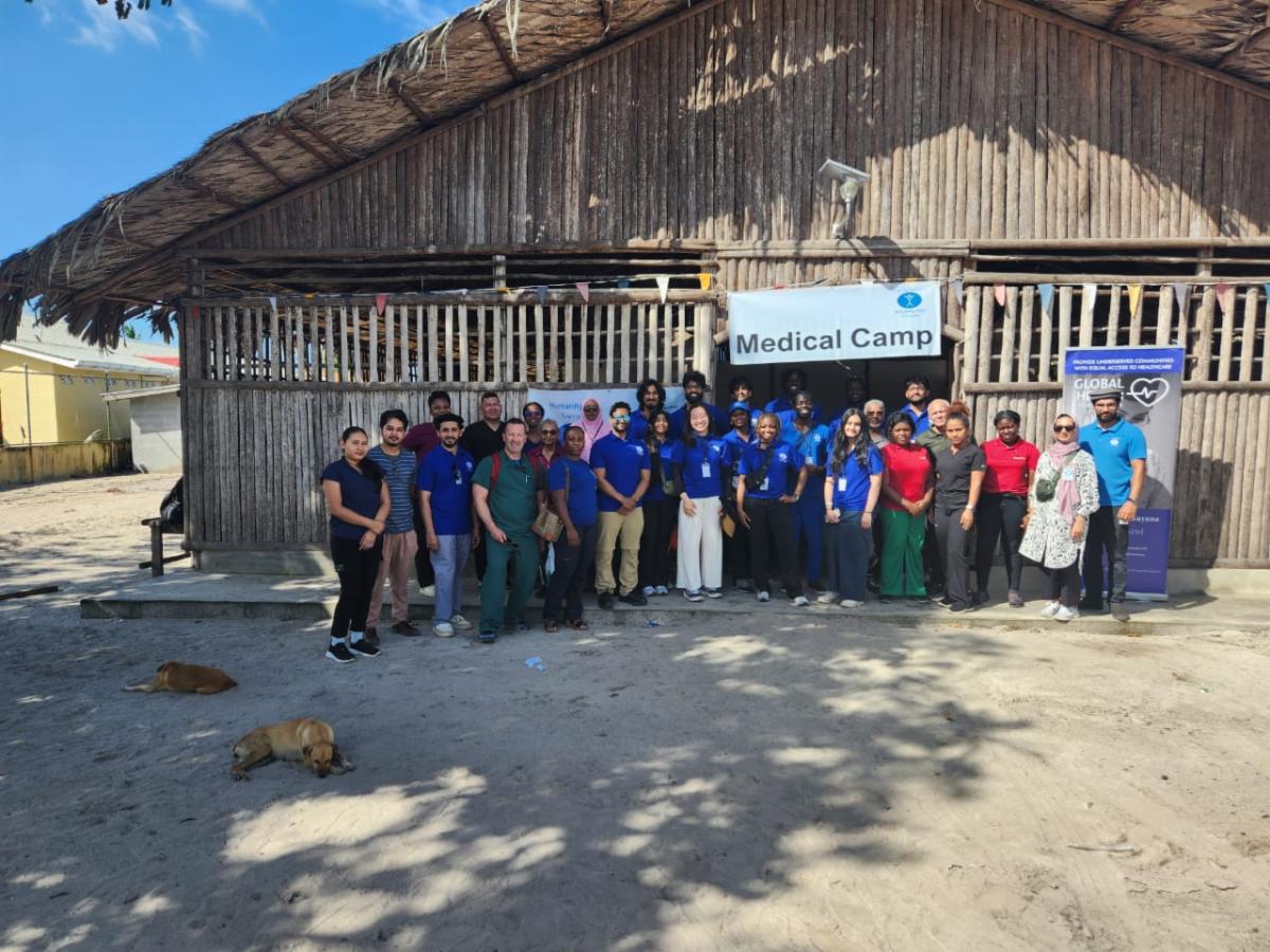 A group photo in front of a building made of wood with a peaked thatched roof. Many members of the group wear matching blue t-shirts. The sign over the door to the building says Medical Camp.