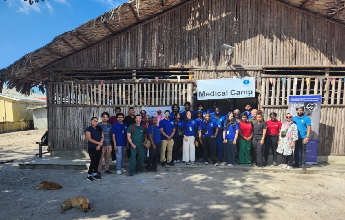 A group photo in front of a building made of wood with a peaked thatched roof. Many members of the group wear matching blue t-shirts. The sign over the door to the building says Medical Camp.