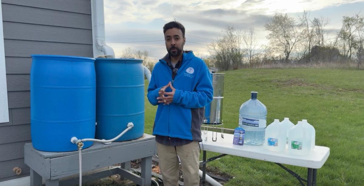 A man stands outside in front of two tables. One of them has two giant tubs of water and the other has multiple sizes of water containers.