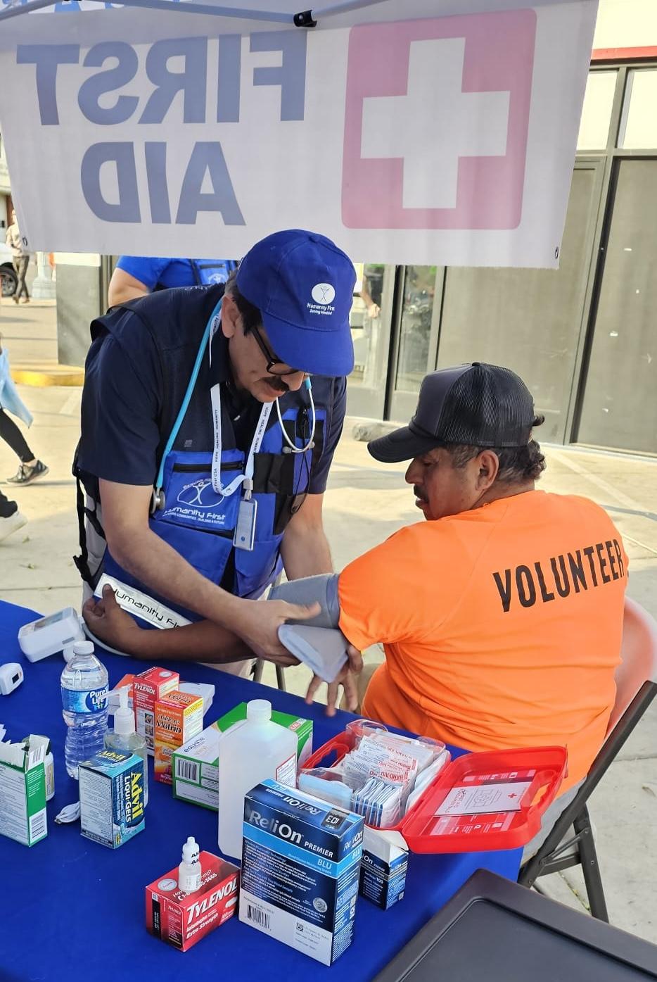 A man wearing blue humanity first gear and a stethoscope puts a blood pressure cuff on a man seated under an open-sided tent. The seated man wears an orange t shirt that says volunteer.