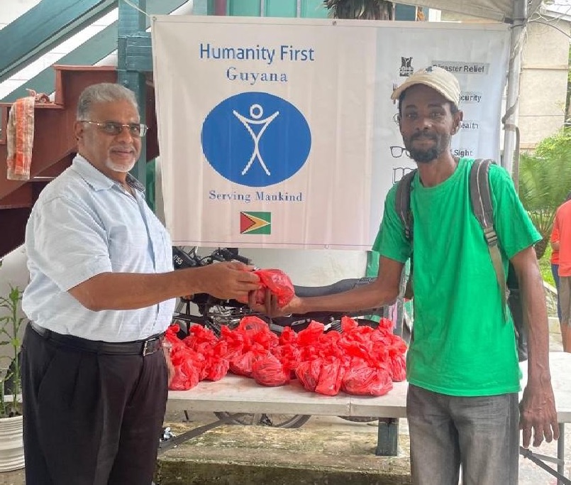 One man hands a red plastic bag of qurbani meat to another man who smiles at the camera. Behind them is a sign for Humanity First Guyana.