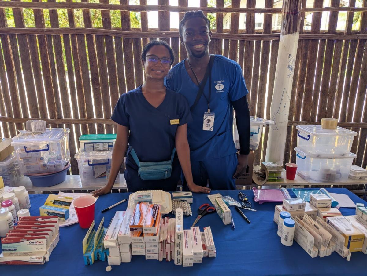 Two people wearing blue medical scrubs stand behind a table full of boxes and bottles of medications.