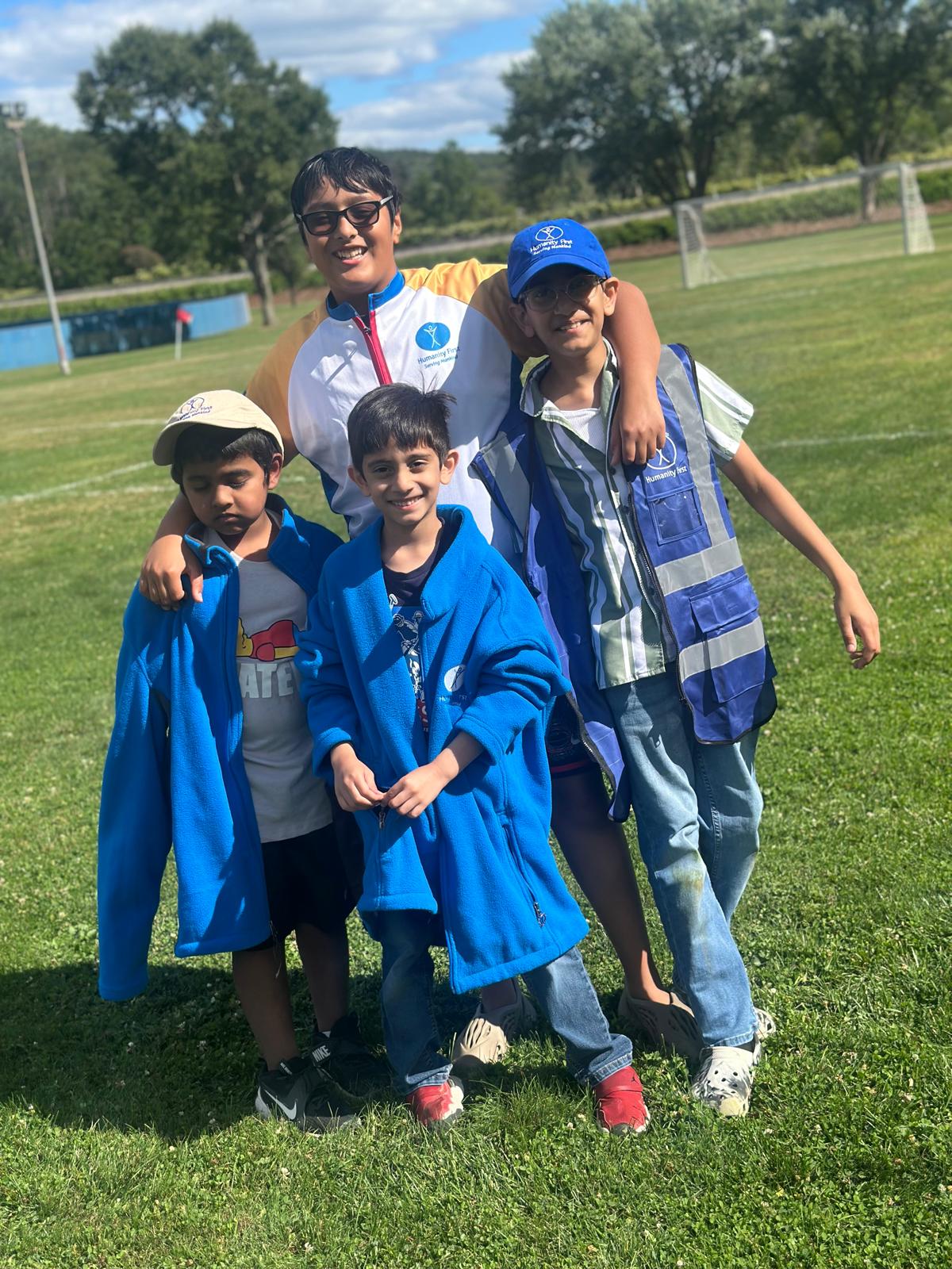 Four young boys wearing blue jackets or humanity first vests pose in a group on a sports field.