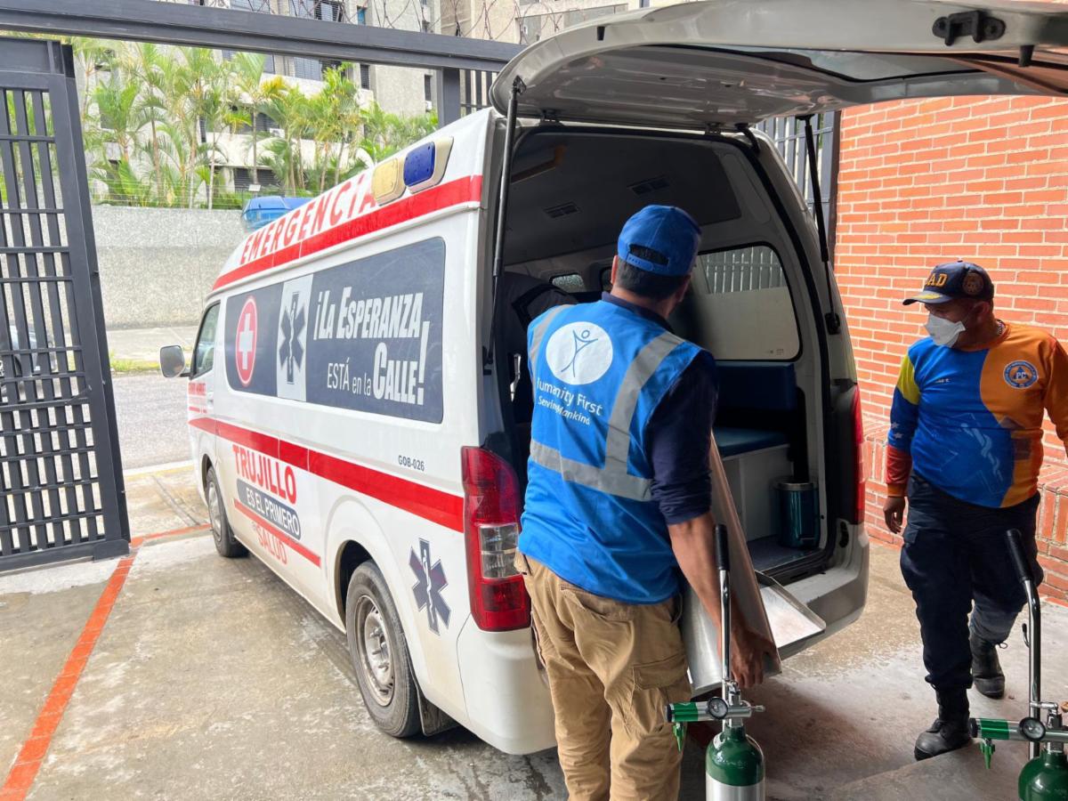 Two men wearing blue Humanity First vests move supplies into the open back of an ambulance.
