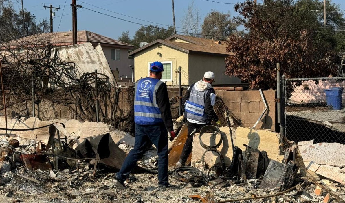 Two men wearing blue vests walk through debris from a burned house