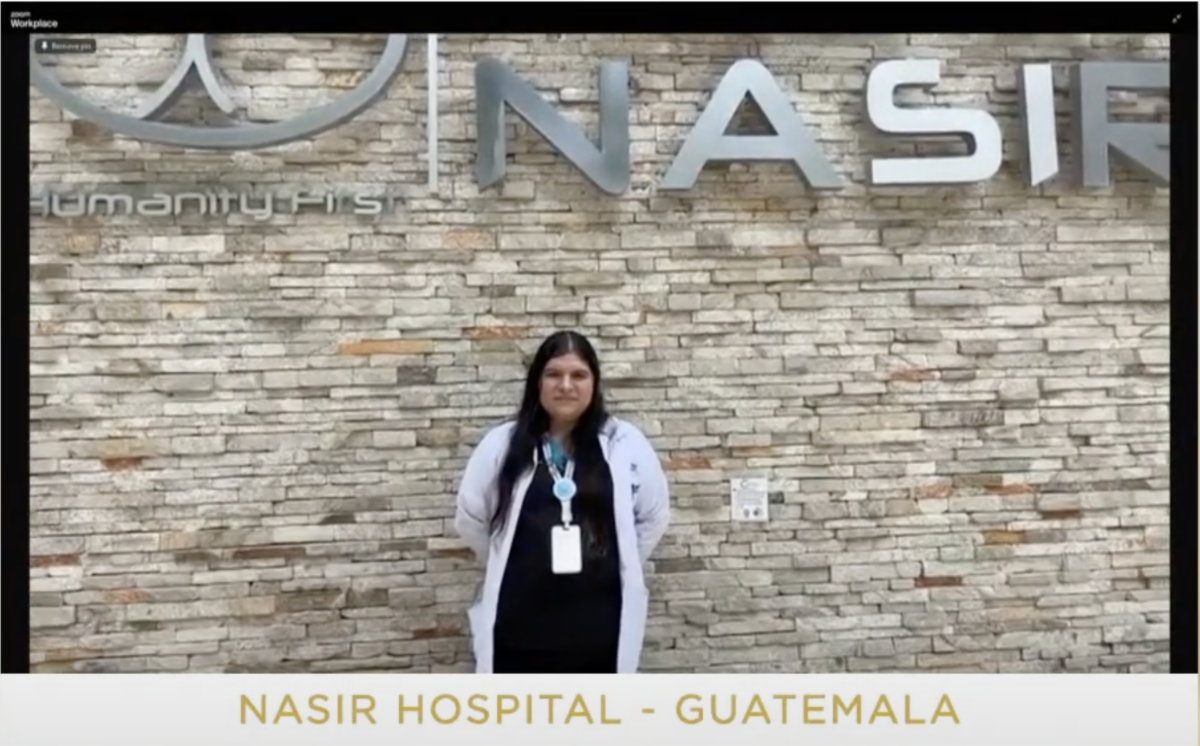 A woman wearing a white medical coat stands in front of the brick outer wall of Nasir Hospital with its sign above her