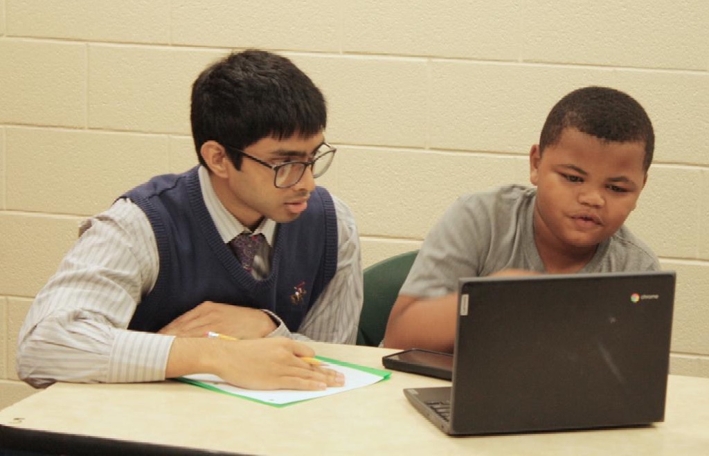 A post-secondary student sits at a table beside a younger student who points at a laptop screen.